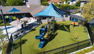 Elevated view of a fenced in play area with play structure and large blue overhead shade incorporated next to a church building with a secondary fenced off play area for younger children.