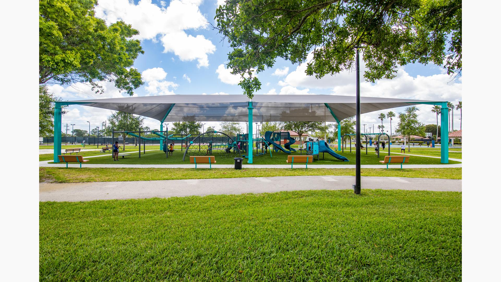 A large grey shade covers a play area with multiple play structures, swing sets, and ziplines at a town park.