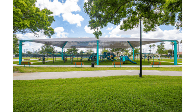 A large grey shade covers a play area with multiple play structures, swing sets, and ziplines at a town park.