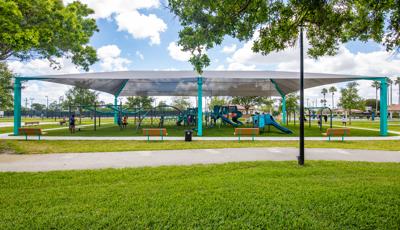 A large grey shade covers a play area with multiple play structures, swing sets, and ziplines at a town park.