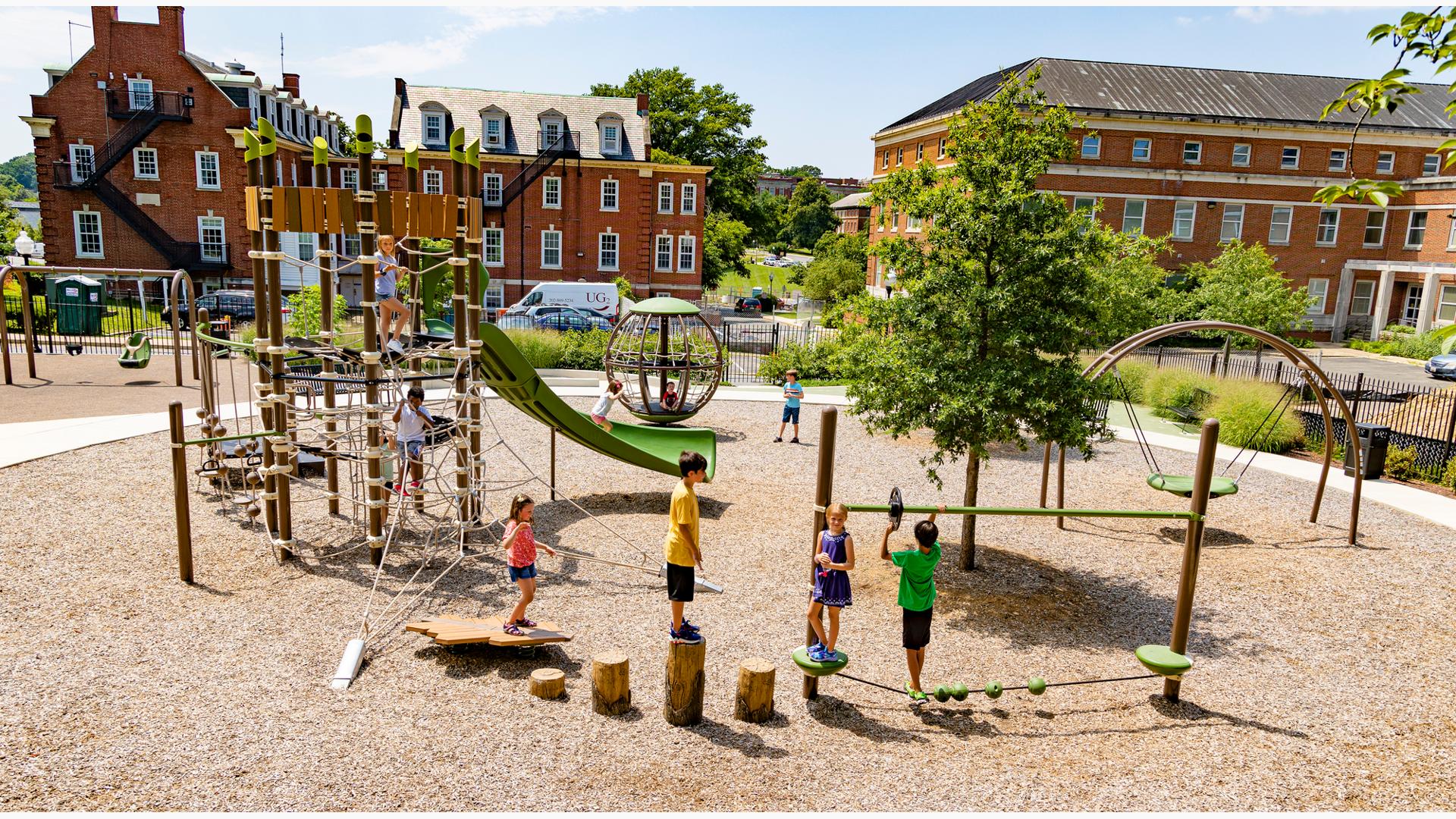 Children play on log foot steppers and a tight rope bridge in the foreground while others climb on a rope climber connected to a larger play structure also made up of climbing ropes. Other play activities surrounding the play structure. The park has a woodchip surfacing and is surrounded by a black iron fence and two large traditional brick buildings. 