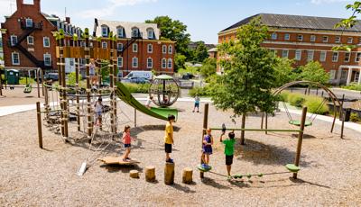 Children play on log foot steppers and a tight rope bridge in the foreground while others climb on a rope climber connected to a larger play structure also made up of climbing ropes. Other play activities surrounding the play structure. The park has a woodchip surfacing and is surrounded by a black iron fence and two large traditional brick buildings. 