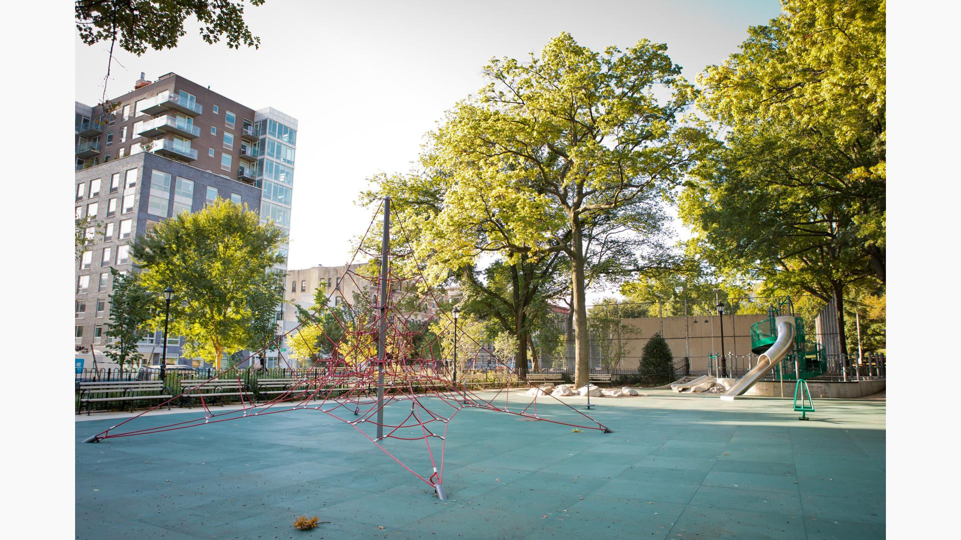 A red roped pyramid shaped climber sits in the foreground as a play structure in the background has a tubular stainless-steel slide. Larger lush trees fill in around the play area of the city park.