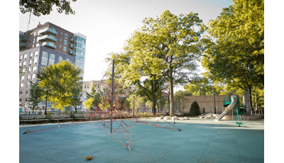 A red roped pyramid shaped climber sits in the foreground as a play structure in the background has a tubular stainless-steel slide. Larger lush trees fill in around the play area of the city park.
