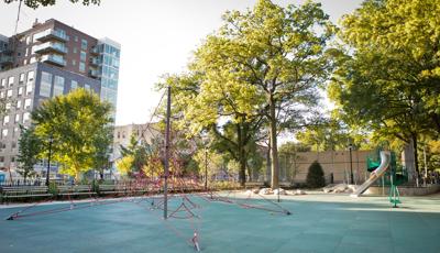 A red roped pyramid shaped climber sits in the foreground as a play structure in the background has a tubular stainless-steel slide. Larger lush trees fill in around the play area of the city park.