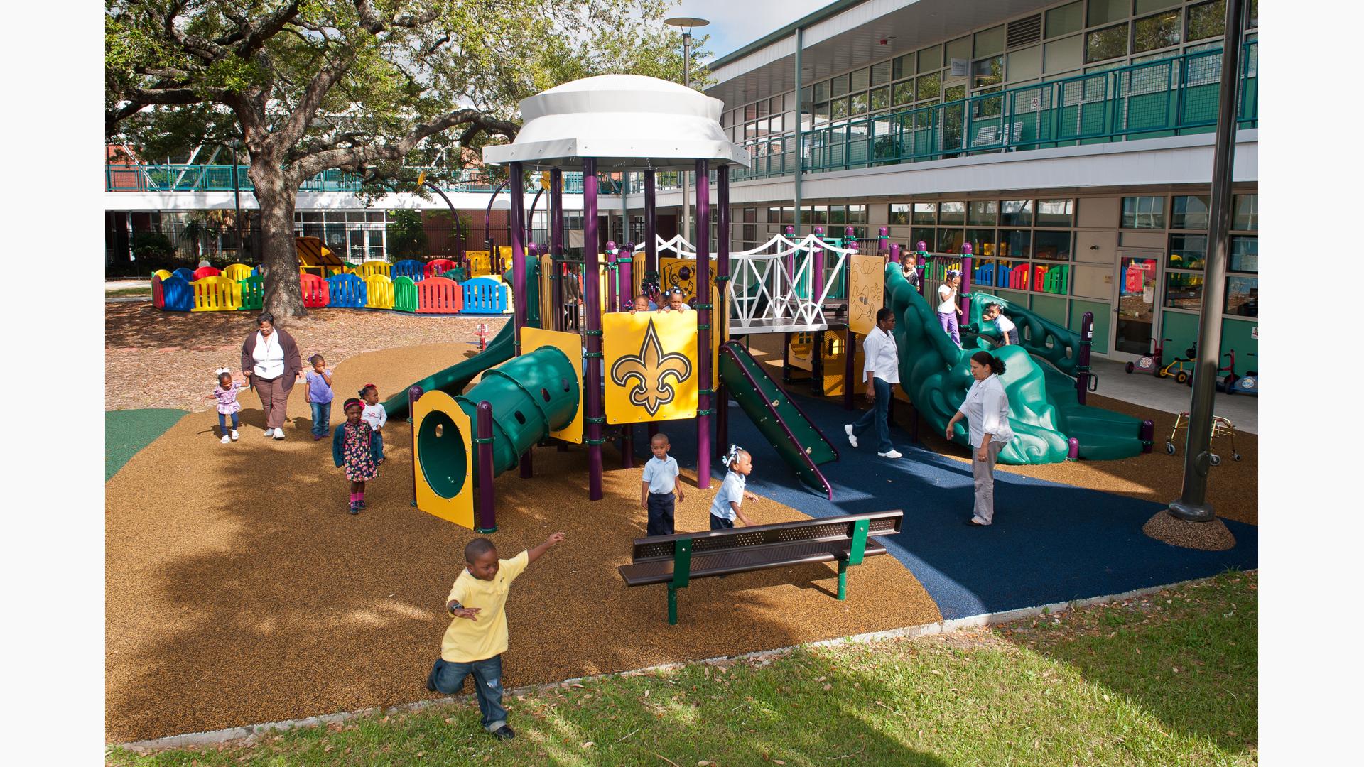 Children play all over a play structure designed in yellow, purple, and greens in the court yard of a large building. One of the play panels has a Fleur-de-lis on it. 