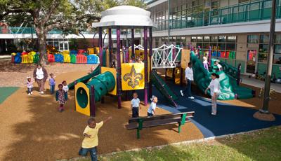 Children play all over a play structure designed in yellow, purple, and greens in the court yard of a large building. One of the play panels has a Fleur-de-lis on it. 