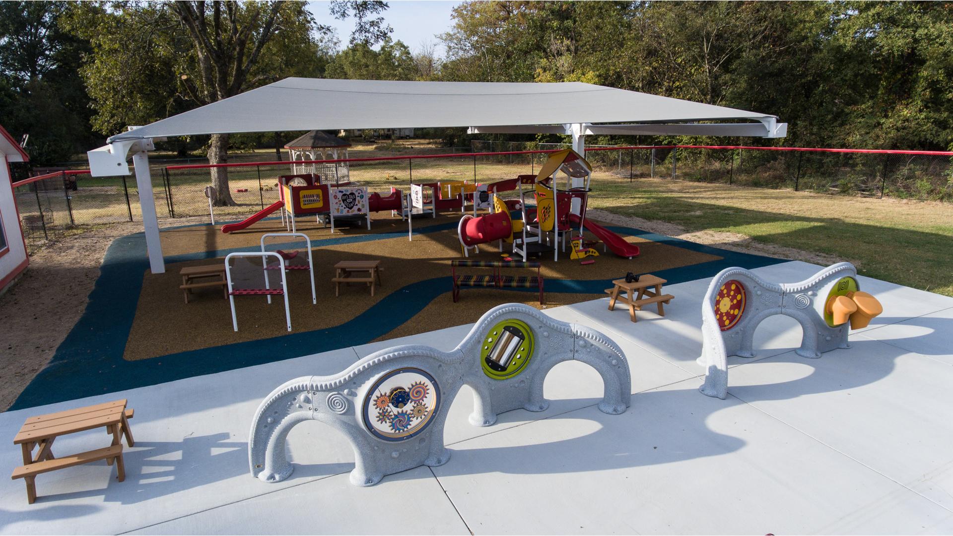Two sensory play panel walls sit in the foreground while in the background a grey shade structure stands over a play area with two play structures and children sized picnic tables. 