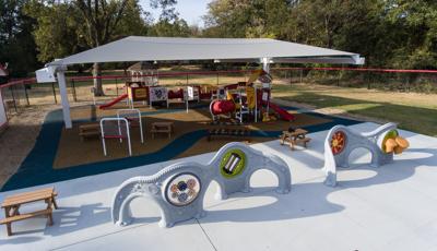 Two sensory play panel walls sit in the foreground while in the background a grey shade structure stands over a play area with two play structures and children sized picnic tables. 