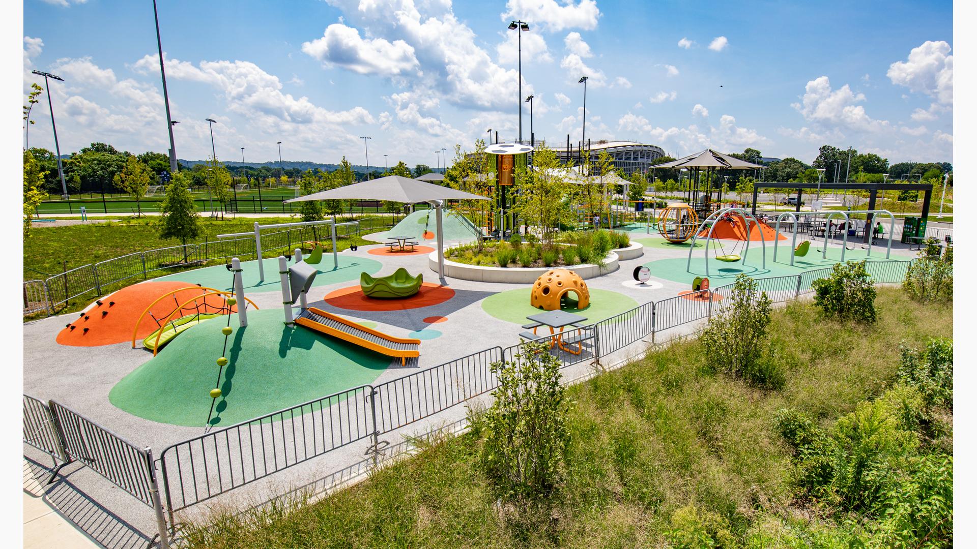 Large play space including merry-go-round, swings, sports-theme playground tower and a playground tower with shade. 