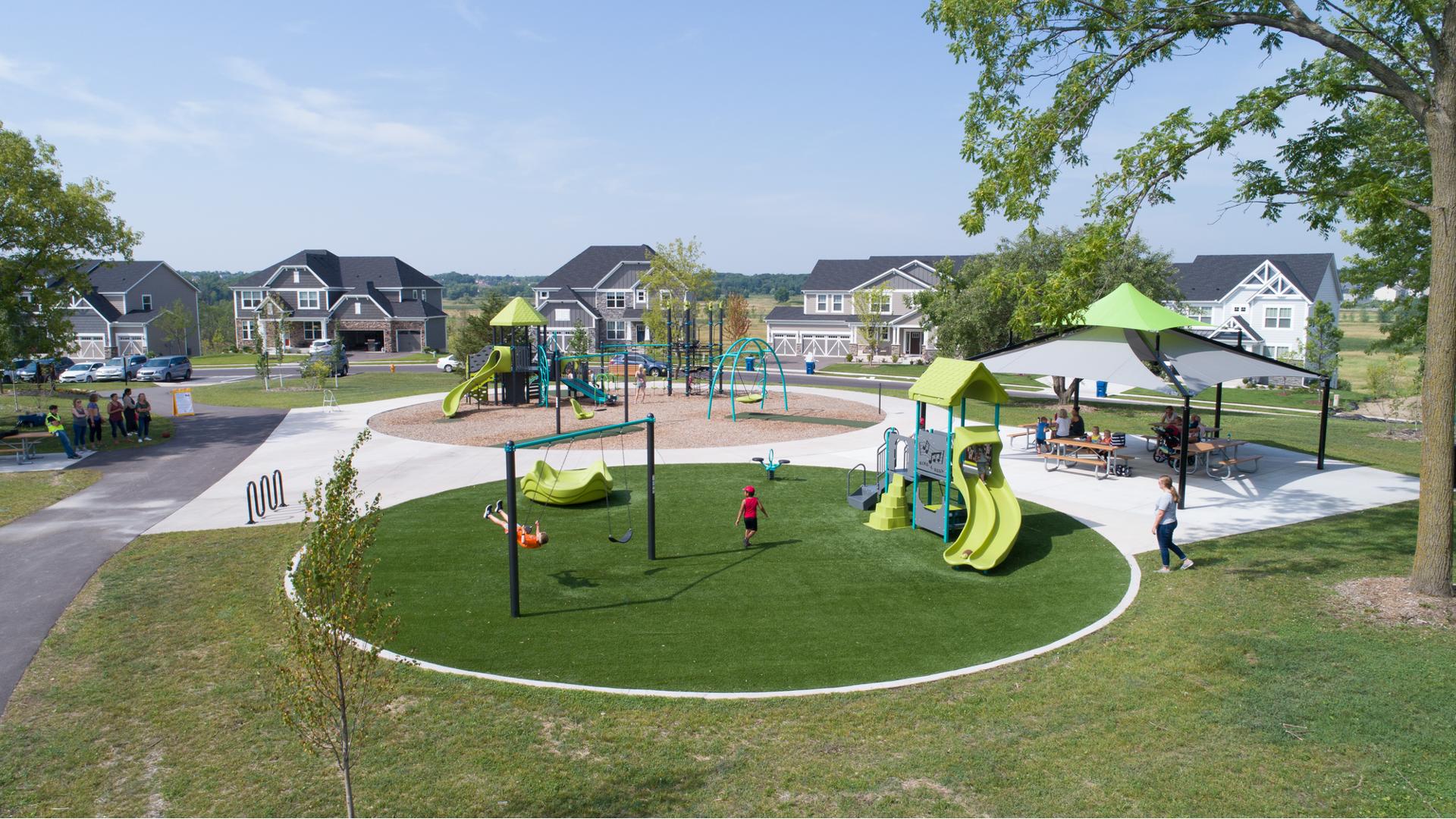 Kids of all ages playing on limon, gray and blue colored playgrounds in a neighborhood park and families gathered under picnic shelter. 