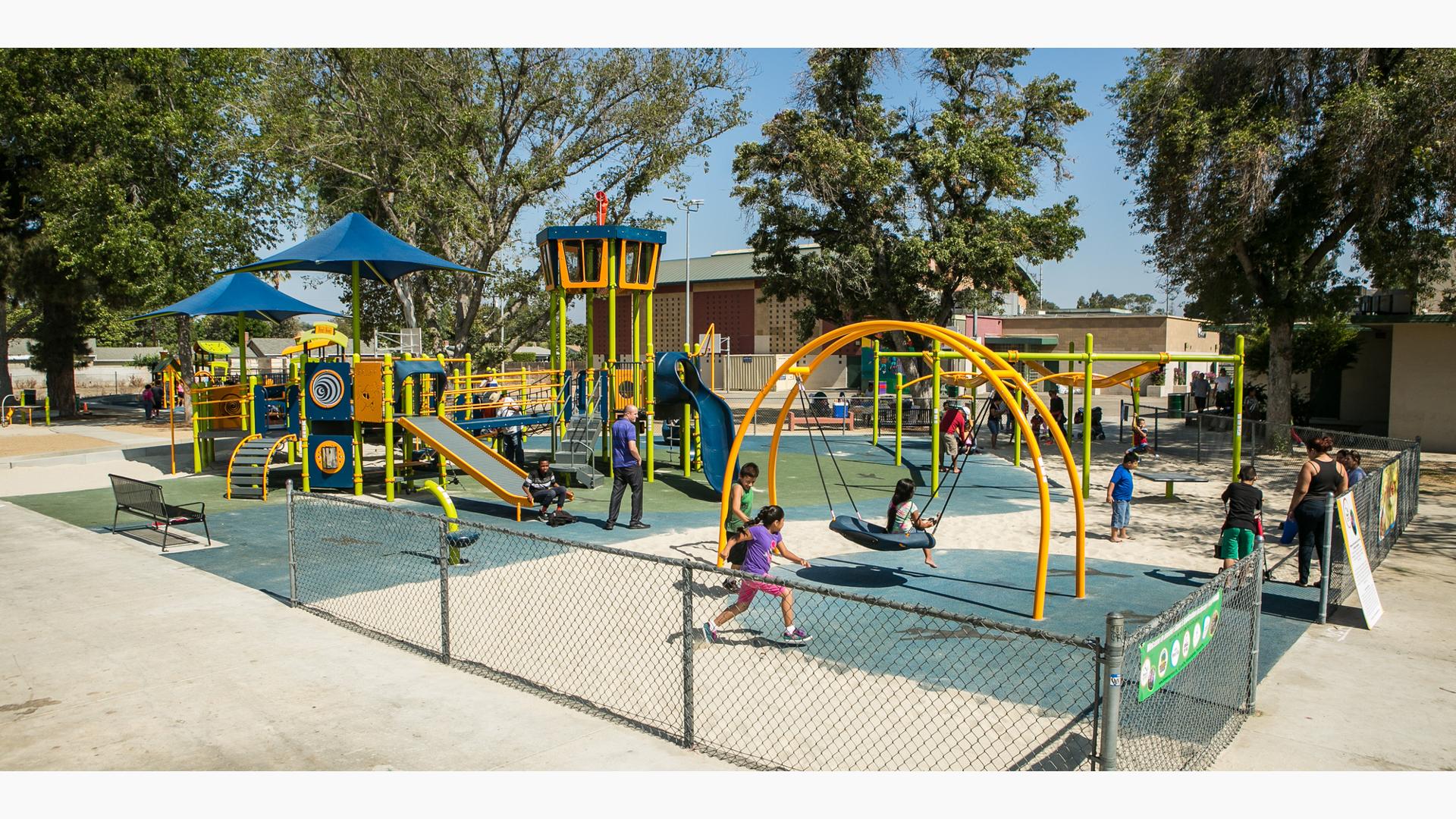 Bright green and orange playground with kids playing. Surfacing is a mix of blue rubber and sand. 