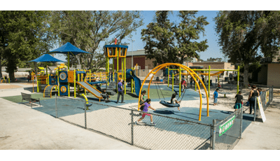 Bright green and orange playground with kids playing. Surfacing is a mix of blue rubber and sand. 