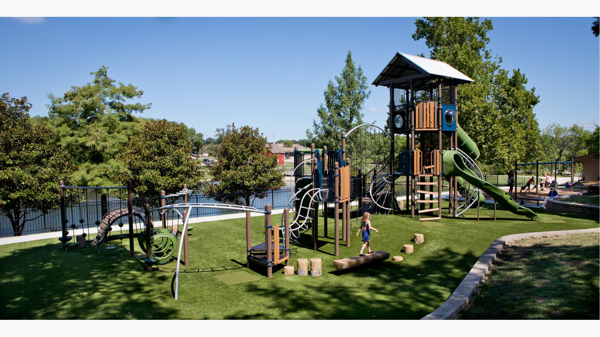 Girl in dress walks on log bench at a playground with green grass surfacing. The playground also includes a tower structure with a nature-inspired look and feel.