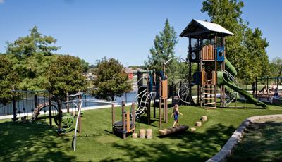 Girl in dress walks on log bench at a playground with green grass surfacing. The playground also includes a tower structure with a nature-inspired look and feel.