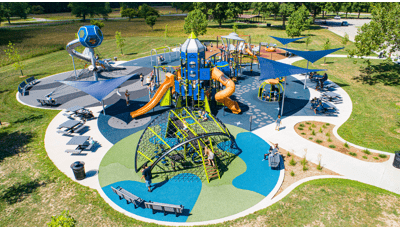 Full elevated view of a space themed playground filled with playing children. The playground safety surfacing is designed like planets, asteroid fields, moons, and stars. A playground tower designed like a rocket and a roof panel like a flying UFO.