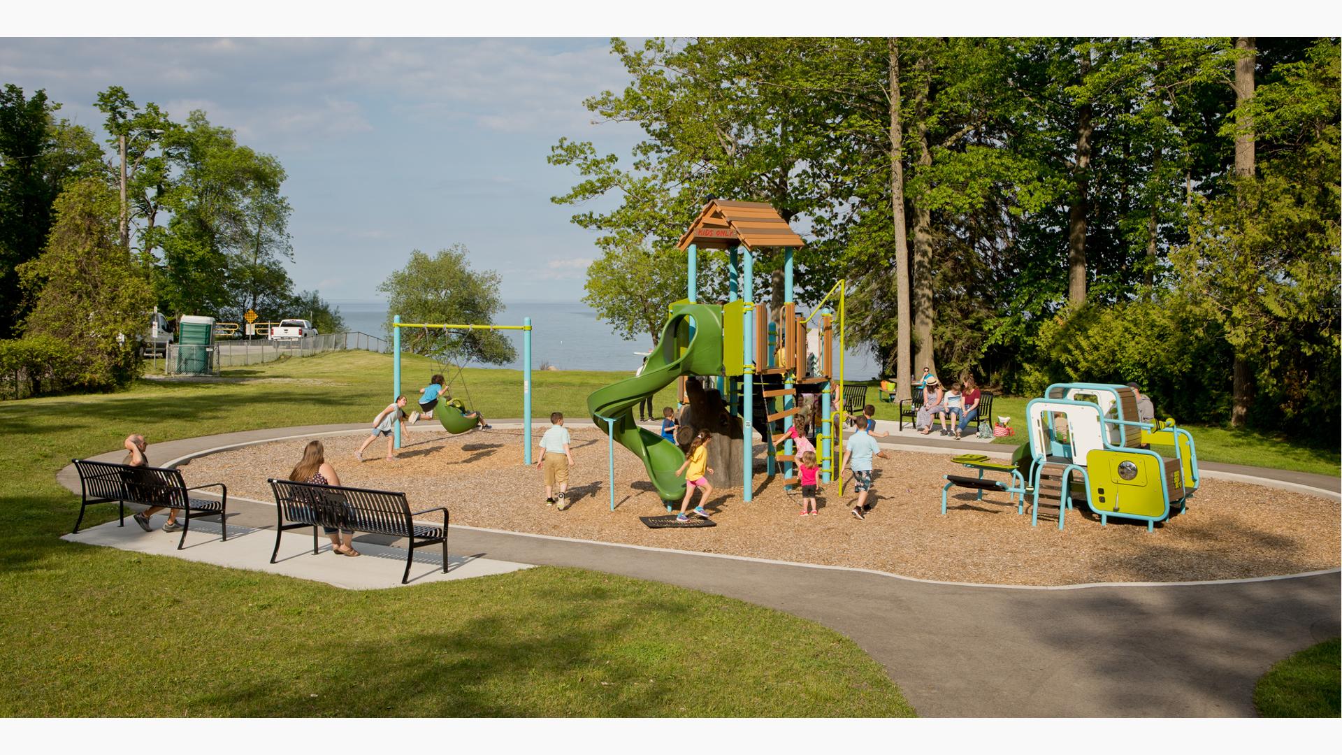 Nature-inspired playground blends into the nearby woods and lake setting of this green and blue playground. Swings located to left of compact playground with woodgrain roof and modern playstructure for toddlers.