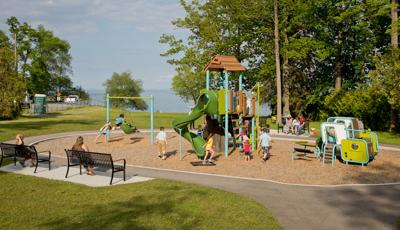 Nature-inspired playground blends into the nearby woods and lake setting of this green and blue playground. Swings located to left of compact playground with woodgrain roof and modern playstructure for toddlers.