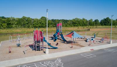 A large play area with woodchip surfacing. Two large tower structures surrounded by other stand alone play activities and multiple swing set bays. A natural grass field fills the background with a lush green tree line while in the foreground a black top is painted with the United States outline and two four square games.