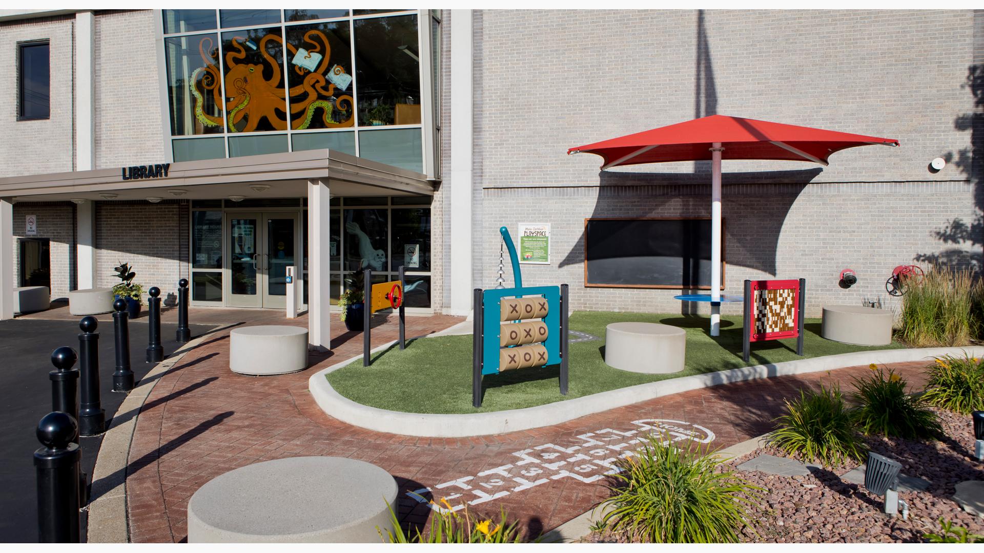 A brick walking path leading into a library building has a hopscotch game painted on it. A turfed area sits to the right of the library entrance with three different play panels, outdoor musical bell instrument, shade station with connected table and a mounted chalk board directly on the buildings outer wall.