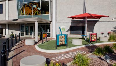 A brick walking path leading into a library building has a hopscotch game painted on it. A turfed area sits to the right of the library entrance with three different play panels, outdoor musical bell instrument, shade station with connected table and a mounted chalk board directly on the buildings outer wall.