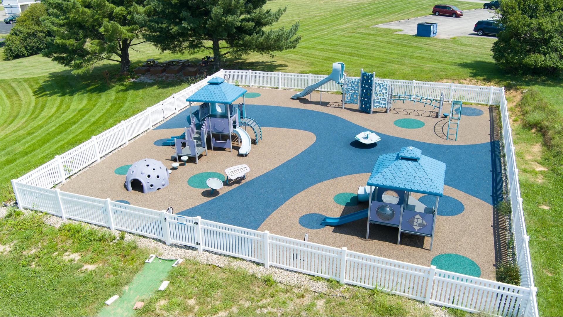Elevated view of a square play space surrounded by a white picket fence with three separate play structures ranging accessibility from toddlers to twelve year old's.