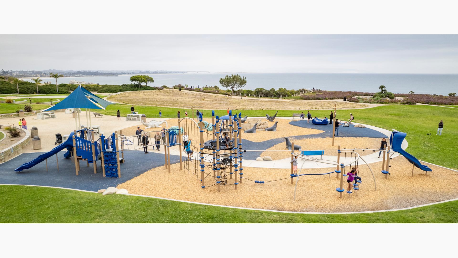 Children play on one long connecting play structure of climbers, slides and bridges with an additional open play space just beyond the play structure all sitting at the top of a bluff overlooking the ocean.