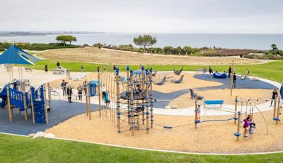 Children play on one long connecting play structure of climbers, slides and bridges with an additional open play space just beyond the play structure all sitting at the top of a bluff overlooking the ocean.