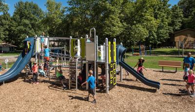 Families playing on park play structure.. A dad holds his kid in his arms as other children climb and run around.