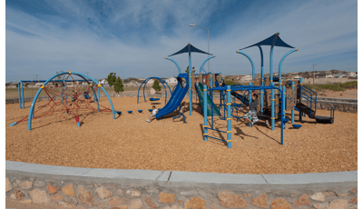 Children play on a park playground with different shades of blue posts, overhead shades, and climbers all surrounded by a neighborhood community. 