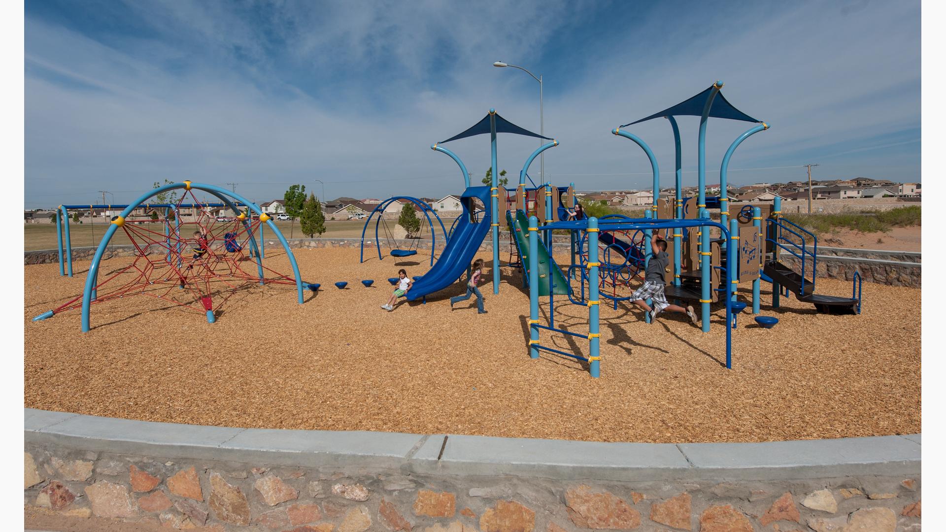Children play on a park playground with different shades of blue posts, overhead shades, and climbers all surrounded by a neighborhood community. 