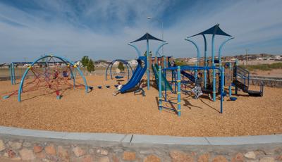 Children play on a park playground with different shades of blue posts, overhead shades, and climbers all surrounded by a neighborhood community. 