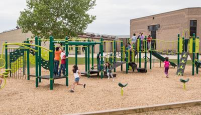 It is a cloudy day as kids at Stanley Knowles School play on the playground. A girl runs to the back of the line for the ZipKruz. Another girl uses the Gyro Twister Spinner. A group of children gather at the top of the slide.