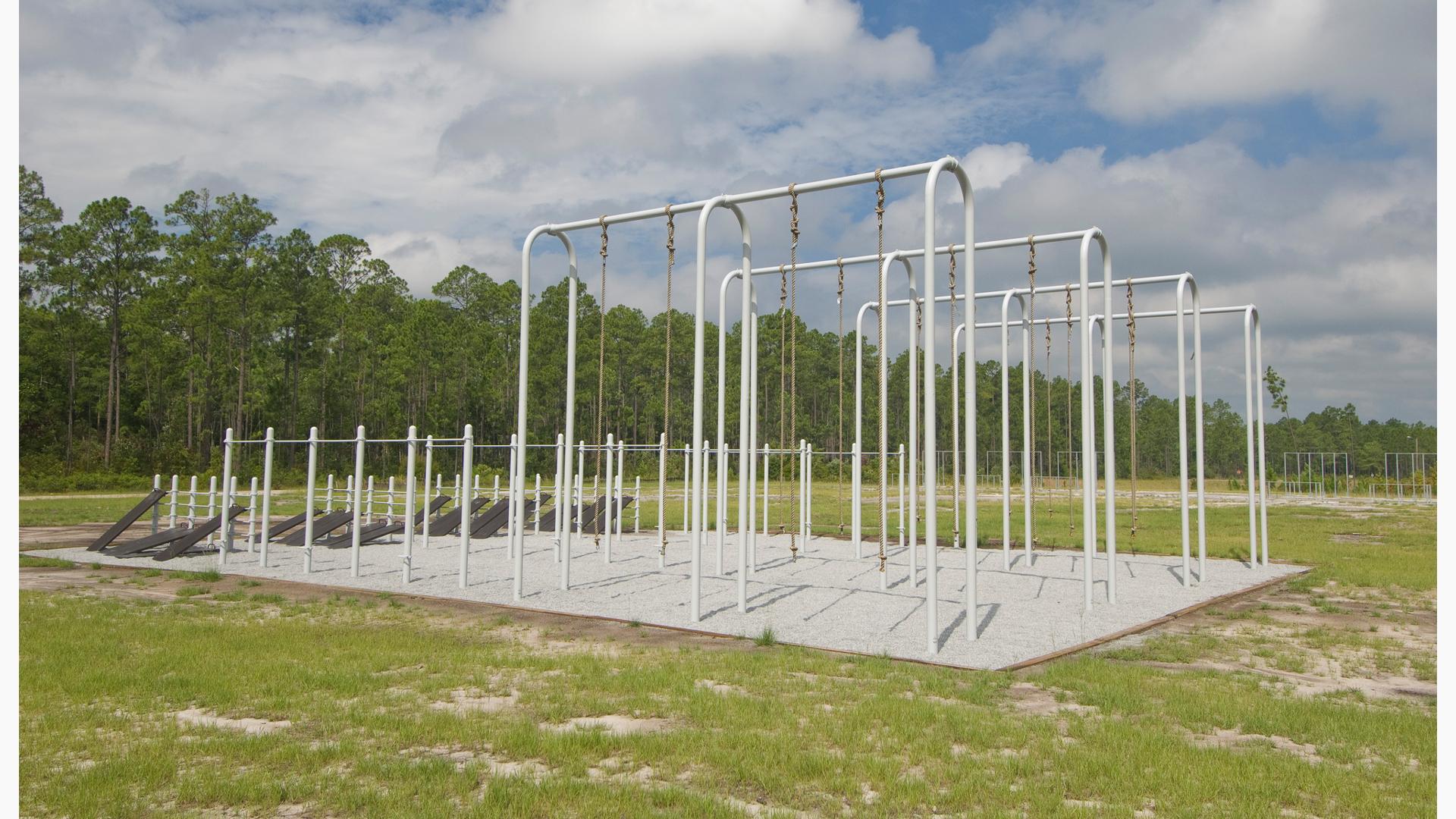 grassy field bordered by trees with rows of military fitness testing equipment. Site includes chin up bars, tall rope climbs and footwork boards.