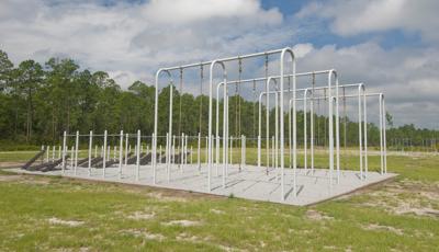 grassy field bordered by trees with rows of military fitness testing equipment. Site includes chin up bars, tall rope climbs and footwork boards.