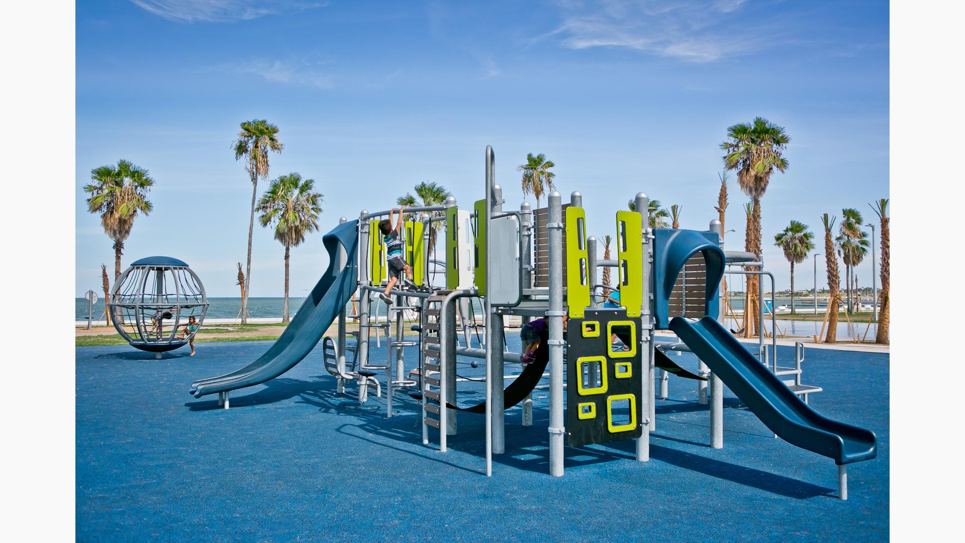Boy climbing on second level of Smart Play structure in McCaughan Park. The park sits by the beach. Palm trees stretch up to the big blue sky.