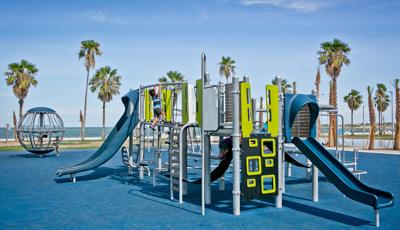 Boy climbing on second level of Smart Play structure in McCaughan Park. The park sits by the beach. Palm trees stretch up to the big blue sky.