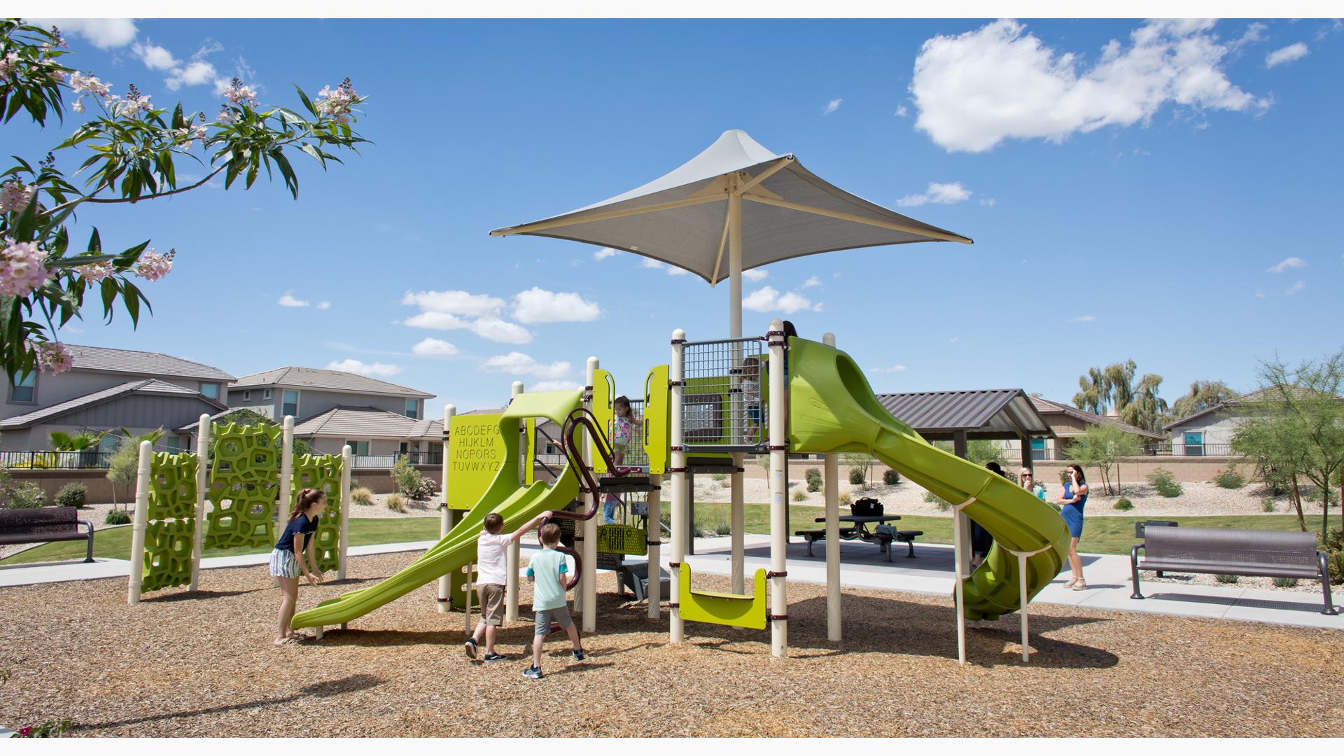 Parents watch their kids have fun on new playground. Two boys climb a logo climber, while an older girl encourages a toddler girl to ride down the double slide.