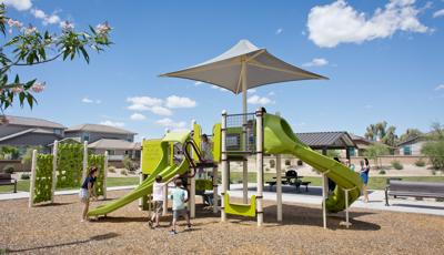 Parents watch their kids have fun on new playground. Two boys climb a logo climber, while an older girl encourages a toddler girl to ride down the double slide.