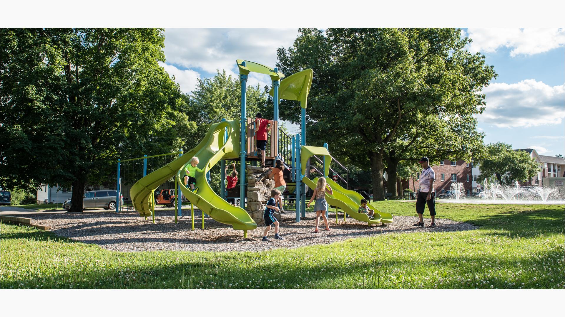 Dad watches his son ride down Double slide as other kids climb rock climber feature. A boy in blue jumps off of Gemini slide.