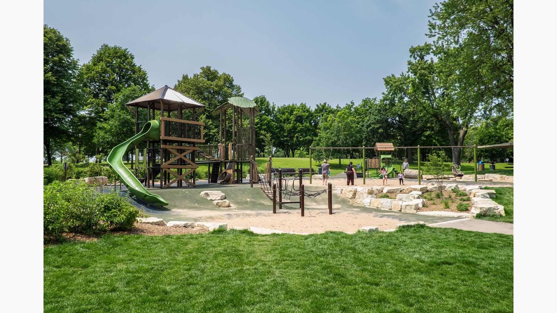 Nature-inspired playground with two towers. One tower has a green slide off the top tier. A smaller playground is in the background along with parent pushing their kids on swings. 