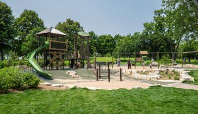 Nature-inspired playground with two towers. One tower has a green slide off the top tier. A smaller playground is in the background along with parent pushing their kids on swings. 