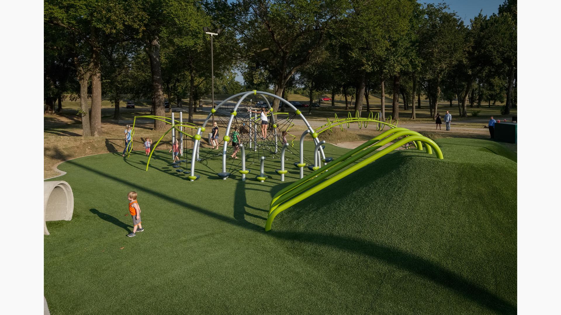 Children climb and play on the ropes of a unique play structure with additional connected spinners, bridges, climbers, and artificial grass underfoot.