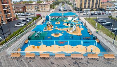 Elevated view of a large play area next to a boardwalk with a ocean theme and two separate play structures and sitting area with large blue square shade structures.