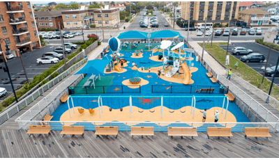 Elevated view of a large play area next to a boardwalk with a ocean theme and two separate play structures and sitting area with large blue square shade structures.