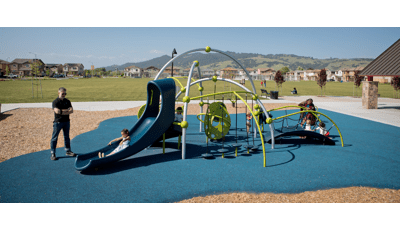 Dad watches daughter ride down slide as toddlers sit on bridge.