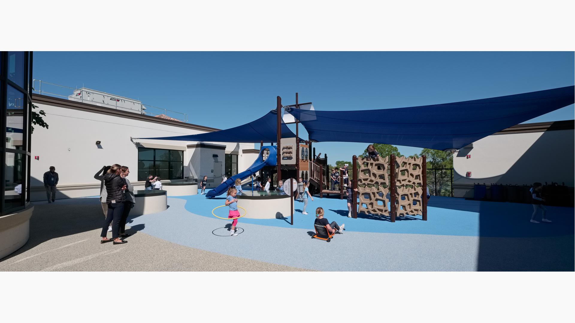 Parents watch as children play on a playground structure shaped like a ship with two large blue shades connecting to the center masts and surrounding school building. 