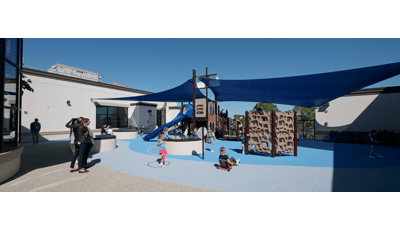 Parents watch as children play on a playground structure shaped like a ship with two large blue shades connecting to the center masts and surrounding school building. 