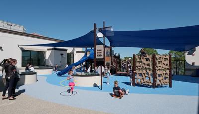 Parents watch as children play on a playground structure shaped like a ship with two large blue shades connecting to the center masts and surrounding school building. 