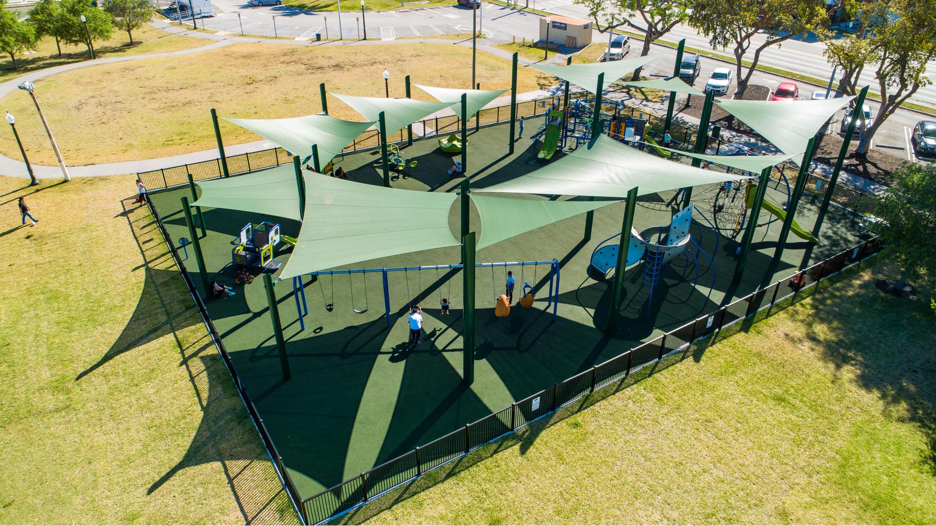 Elevated view of a large fenced in play area with multiple play structures, climbers and swing sets all covered by large green shade sails overhead.